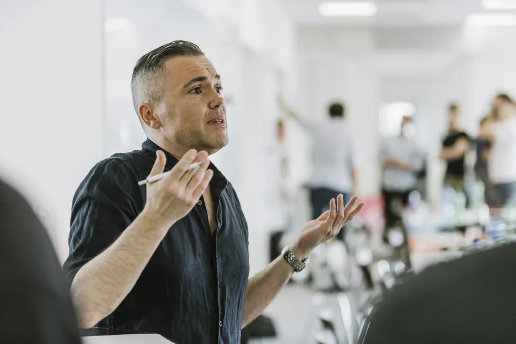Eine Person in einem dunklen Hemd gestikuliert mit den Händen, während sie in einem hellen Raum spricht. In einer Hand hält sie einen Stift. Im Hintergrund sind mehrere Personen und Bürogeräte verschwommen. Eine Person in einem dunklen Hemd gestikuliert mit den Händen, während sie in einem hellen Raum spricht. In einer Hand hält sie einen Stift. Im Hintergrund sind mehrere Personen und Bürogeräte verschwommen.