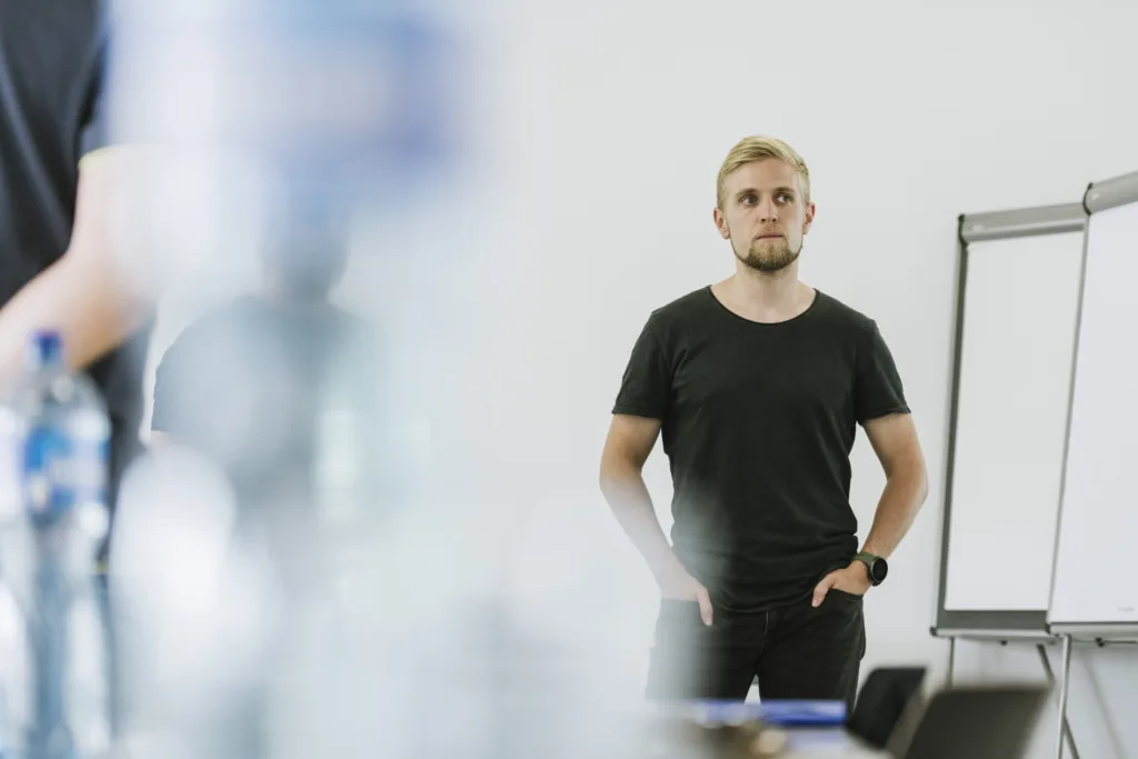 Ein Mann mit blonden Haaren, einem schwarzen T-Shirt und Jeans steht mit den Händen in den Taschen in einem Raum, hinter dem sich leere Flipcharts befinden und im Vordergrund unscharfe Flaschen zu sehen sind. Ein Mann mit blonden Haaren, einem schwarzen T-Shirt und Jeans steht mit den Händen in den Taschen in einem Raum, hinter dem sich leere Flipcharts befinden und im Vordergrund unscharfe Flaschen zu sehen sind.