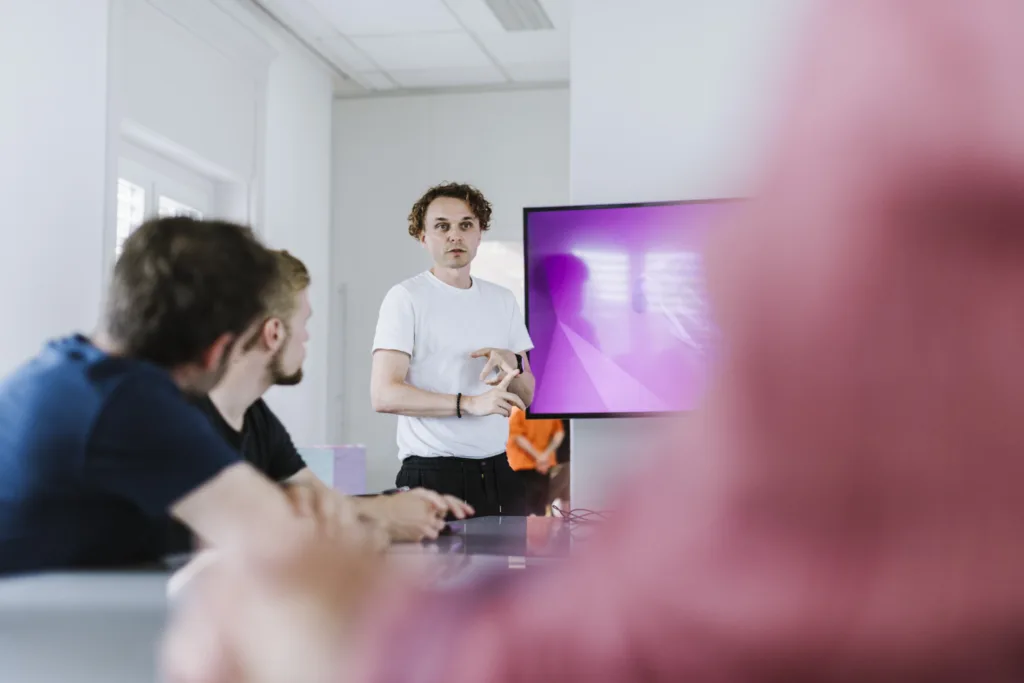 Eine Person im weißen Hemd steht vor einem Bildschirm und hält eine Präsentation. Im Vordergrund sind zwei sitzende Personen zu sehen, die aufmerksam zuhören (teilweise unscharf). Der Raum ist hell, da natürliches Licht durch die Fenster fällt. Eine Person im weißen Hemd steht vor einem Bildschirm und hält eine Präsentation. Im Vordergrund sind zwei sitzende Personen zu sehen, die aufmerksam zuhören (teilweise unscharf). Der Raum ist hell, da natürliches Licht durch die Fenster fällt.