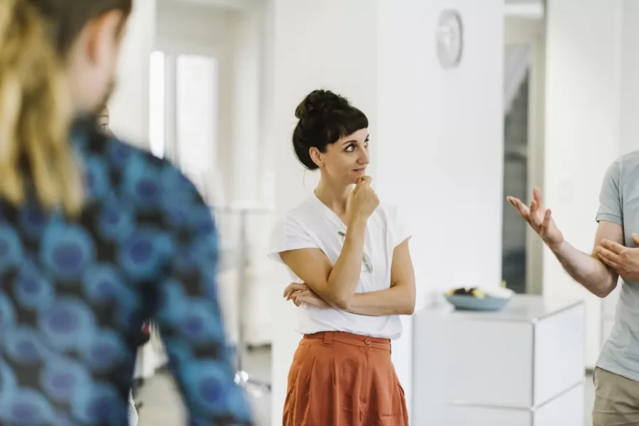 A woman in a white shirt and orange skirt stands thoughtfully with her hand on her chin. She is listening to a person gesturing in front of her in a bright room with white walls and a clock.