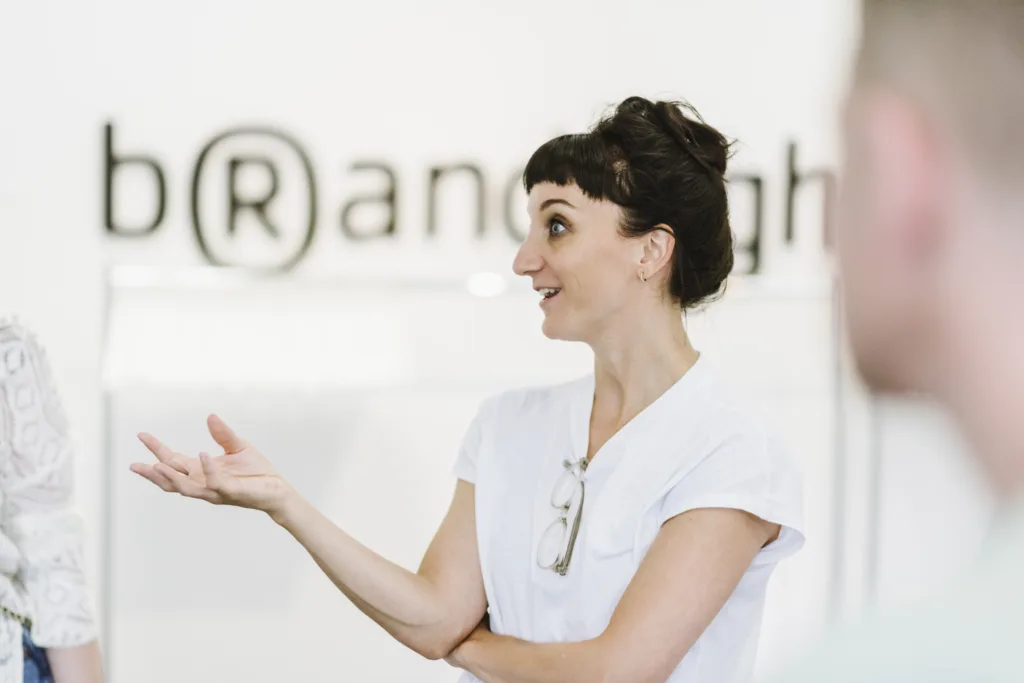 Eine Frau mit kurzen dunklen Haaren und einem weißen Hemd spricht angeregt. Auf ihrem Hemd liegt eine Brille. Der Hintergrund ist verschwommen und zeigt das Wort „Brand“ teilweise mit dem Buchstaben „R“ in Klammern. Eine weitere Person ist teilweise sichtbar. Eine Frau mit kurzen dunklen Haaren und einem weißen Hemd spricht angeregt. Auf ihrem Hemd liegt eine Brille. Der Hintergrund ist verschwommen und zeigt das Wort „Brand“ teilweise mit dem Buchstaben „R“ in Klammern. Eine weitere Person ist teilweise sichtbar.
