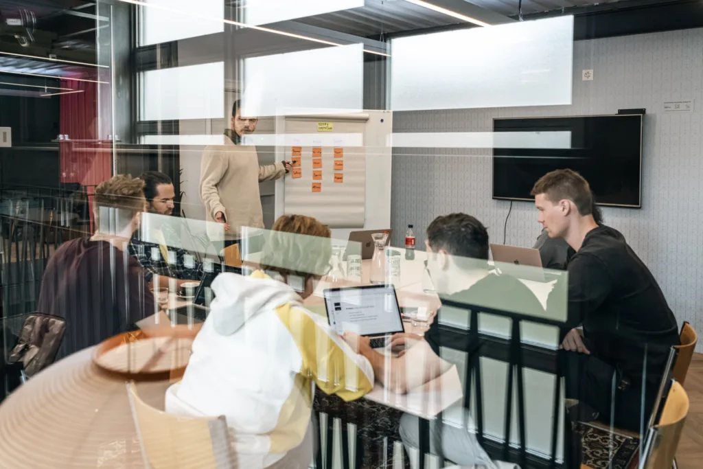 Eine Gruppe von Menschen sitzt um einen Konferenztisch in einem Büro. Eine Person steht an einem Flipchart und hält eine Präsentation. Laptops und Papiere liegen auf dem Tisch. Der Raum hat große Fenster, durch die sich das Innere spiegelt, und einen an der Wand montierten Bildschirm. Eine Gruppe von Menschen sitzt um einen Konferenztisch in einem Büro. Eine Person steht an einem Flipchart und hält eine Präsentation. Laptops und Papiere liegen auf dem Tisch. Der Raum hat große Fenster, durch die sich das Innere spiegelt, und einen an der Wand montierten Bildschirm.