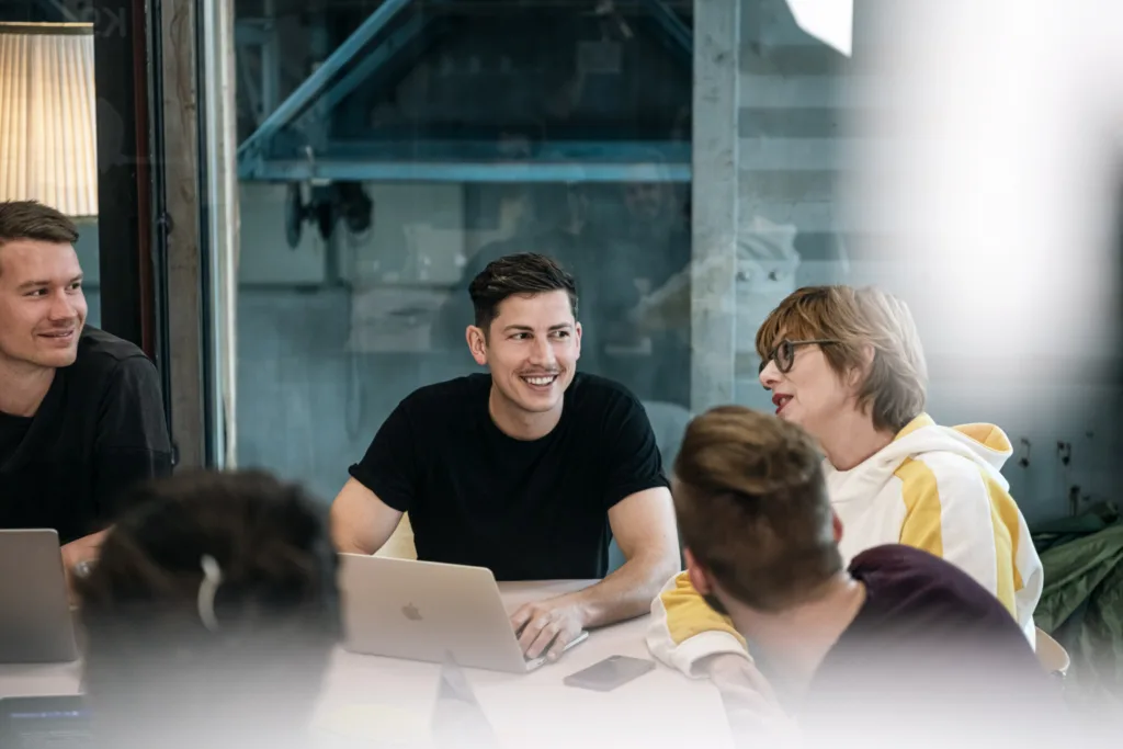 Eine Gruppe von Menschen sitzt mit Laptops um einen Tisch und unterhält sich. Eine Person lächelt eine andere an, die gerade spricht. Die Umgebung wirkt locker und kooperativ, es herrscht eine entspannte Atmosphäre. Eine Gruppe von Menschen sitzt mit Laptops um einen Tisch und unterhält sich. Eine Person lächelt eine andere an, die gerade spricht. Die Umgebung wirkt locker und kooperativ, es herrscht eine entspannte Atmosphäre.