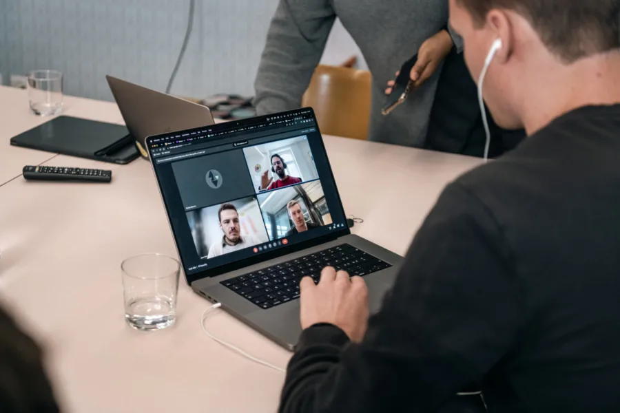 A person wearing earphones sits at a table, using a laptop for a video call with four people visible on screen. Another person stands nearby holding a smartphone. The table also has a glass and another open laptop.