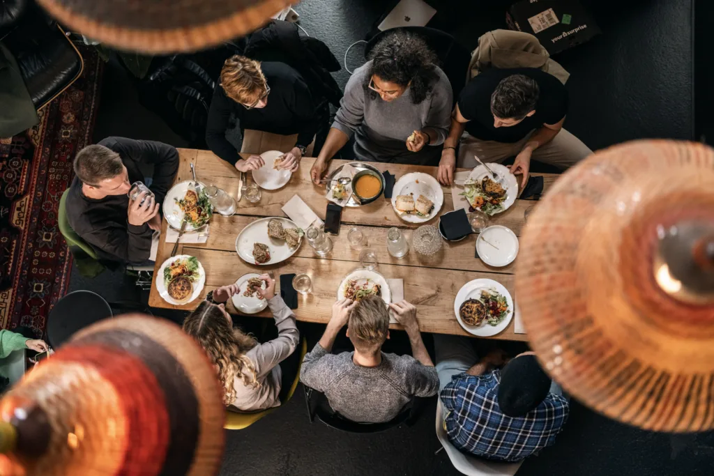 Eine Gruppe von Menschen sitzt um einen Holztisch und isst gemeinsam. Auf dem Tisch stehen Teller mit Essen und Getränken. Von oben betrachtet wirkt die Umgebung warm und einladend, und Hängelampen sorgen für eine gemütliche Atmosphäre. Eine Gruppe von Menschen sitzt um einen Holztisch und isst gemeinsam. Auf dem Tisch stehen Teller mit Essen und Getränken. Von oben betrachtet wirkt die Umgebung warm und einladend, und Hängelampen sorgen für eine gemütliche Atmosphäre.