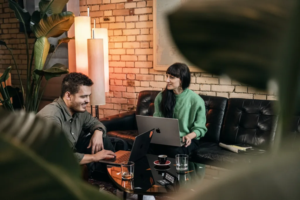 Zwei Personen sitzen auf einem schwarzen Ledersofa und arbeiten an Laptops. Ein Mann mit kurzen Haaren lächelt, während eine Frau mit langen dunklen Haaren einen grünen Pullover trägt. Im Hintergrund sind eine große Lampe und eine Backsteinmauer zu sehen, Pflanzen rahmen die Szene ein. Zwei Personen sitzen auf einem schwarzen Ledersofa und arbeiten an Laptops. Ein Mann mit kurzen Haaren lächelt, während eine Frau mit langen dunklen Haaren einen grünen Pullover trägt. Im Hintergrund sind eine große Lampe und eine Backsteinmauer zu sehen, Pflanzen rahmen die Szene ein.