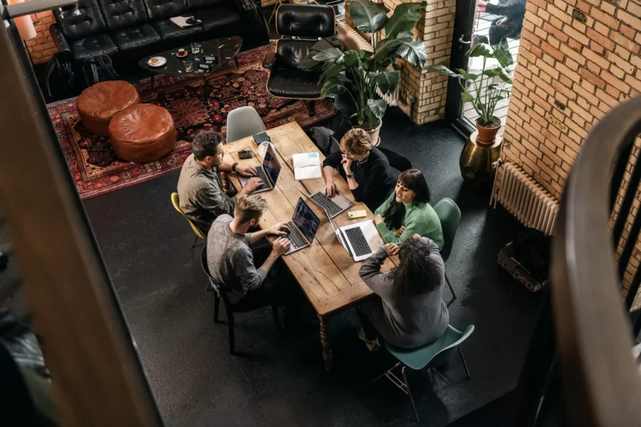 Five people sit around a wooden table working on laptops in a cozy room with a rug, leather couches, and large plants. The setting has brick walls and a warm atmosphere, viewed from above.