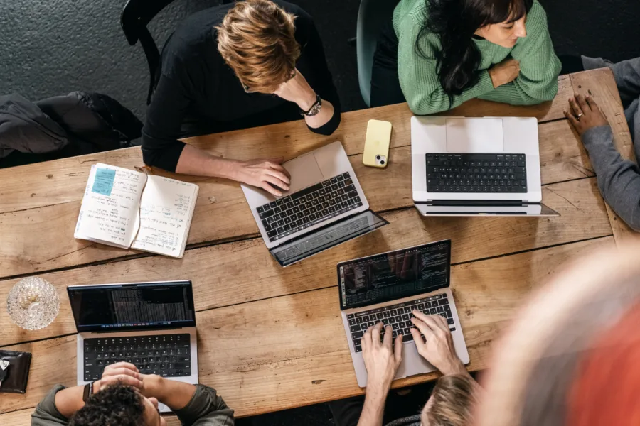 A group of people sit around a wooden table working on laptops. Notebooks and a smartphone are scattered on the table. One person wears a green sweater and others have casual clothing. The setting appears to be a collaborative workspace.