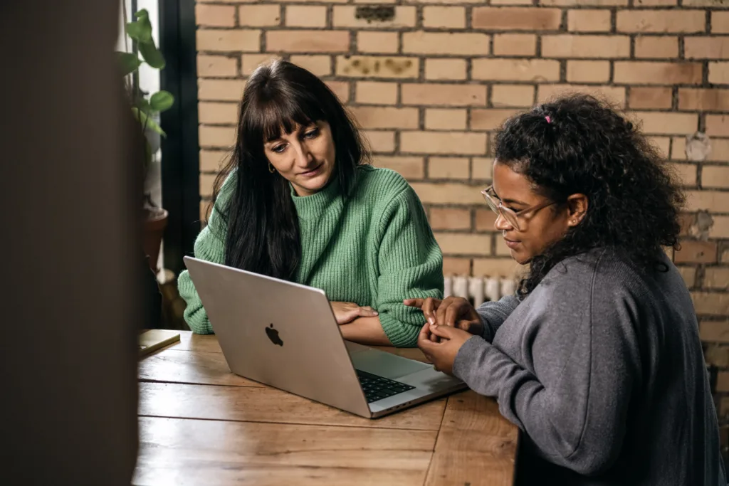 Zwei Frauen sitzen mit einem Laptop an einem Holztisch. Die eine trägt einen grünen Pullover, die andere einen grauen. Sie unterhalten sich, wobei die Frau im grauen Pullover mit den Händen gestikuliert. Im Hintergrund sind eine Backsteinmauer und Pflanzen zu sehen. Zwei Frauen sitzen mit einem Laptop an einem Holztisch. Die eine trägt einen grünen Pullover, die andere einen grauen. Sie unterhalten sich, wobei die Frau im grauen Pullover mit den Händen gestikuliert. Im Hintergrund sind eine Backsteinmauer und Pflanzen zu sehen.
