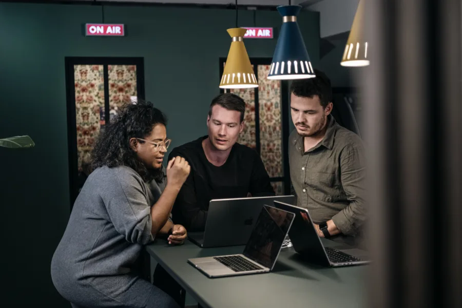 Three people are gathered around a table with laptops, engaged in discussion. They are in a room with ON AIR signs lit up above them, and various pendant lights hang from the ceiling.