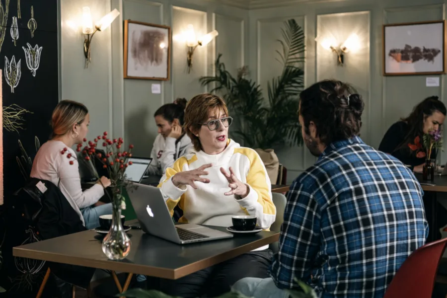 People are sitting in a cozy cafe with wooden walls, engaging in conversations. A woman in a yellow and white sweater talks to a man in a plaid shirt at a table with a laptop. Others work on laptops with plants and artwork nearby.