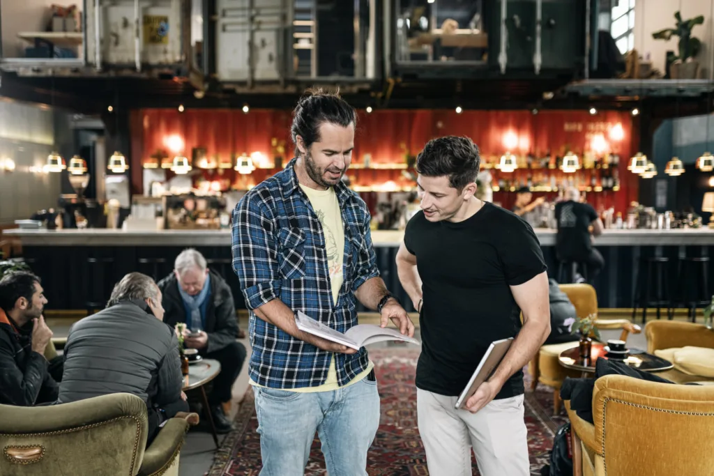 Zwei Männer unterhalten sich und lesen ein Buch in einem gemütlichen Café mit warmer Beleuchtung. Einer trägt ein kariertes Hemd und Jeans, der andere ein schwarzes T-Shirt. Im Hintergrund sitzen Leute an Tischen und ein Barbereich ist sichtbar. Zwei Männer unterhalten sich und lesen ein Buch in einem gemütlichen Café mit warmer Beleuchtung. Einer trägt ein kariertes Hemd und Jeans, der andere ein schwarzes T-Shirt. Im Hintergrund sitzen Leute an Tischen und ein Barbereich ist sichtbar.