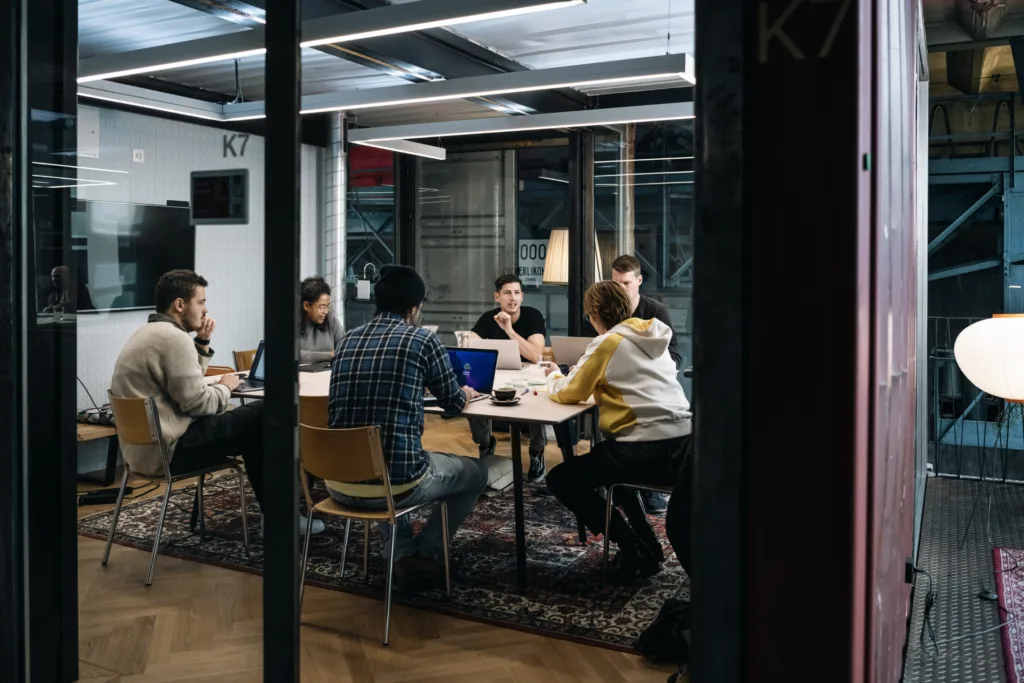 Eine Gruppe von Menschen sitzt in einem modernen Büro um einen Konferenztisch und diskutiert. Einige haben Laptops aufgeklappt und an der Wand hängt ein großer Bildschirm. Der Raum ist von Glaswänden umgeben und unter dem Tisch liegt ein Teppich. Eine Gruppe von Menschen sitzt in einem modernen Büro um einen Konferenztisch und diskutiert. Einige haben Laptops aufgeklappt und an der Wand hängt ein großer Bildschirm. Der Raum ist von Glaswänden umgeben und unter dem Tisch liegt ein Teppich.