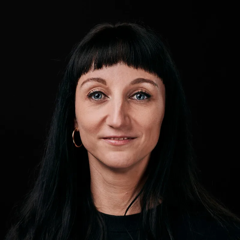 A person with long dark hair and bangs faces the camera against a dark background, embodying a strong sense of individuality within their team. They wear a black top and gold hoop earrings, their expression neutral but subtly commanding.