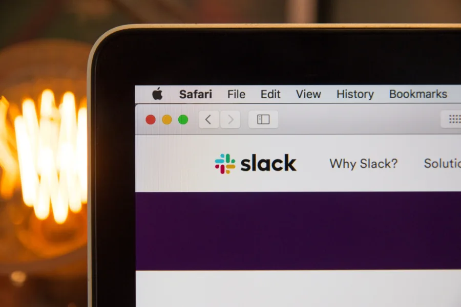 Eine Nahaufnahme zeigt einen Computerbildschirm mit der im Safari-Browser geöffneten Slack-Website, komplett mit Browser-Steuerelementen oben links. Das bekannte Slack-Logo und der Text fallen auf, während im Hintergrund eine verschwommene Glühbirne sanft leuchtet.