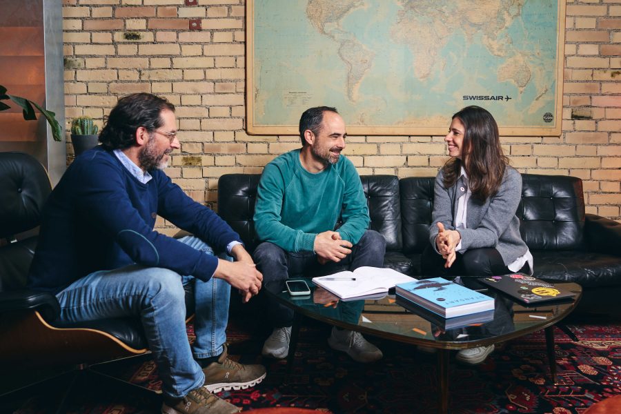 Three people sit on black sofas around a glass coffee table with books and papers, smiling and talking about digital solutions. Behind them is a brick wall with a large world map, creating a relaxed and informal setting.