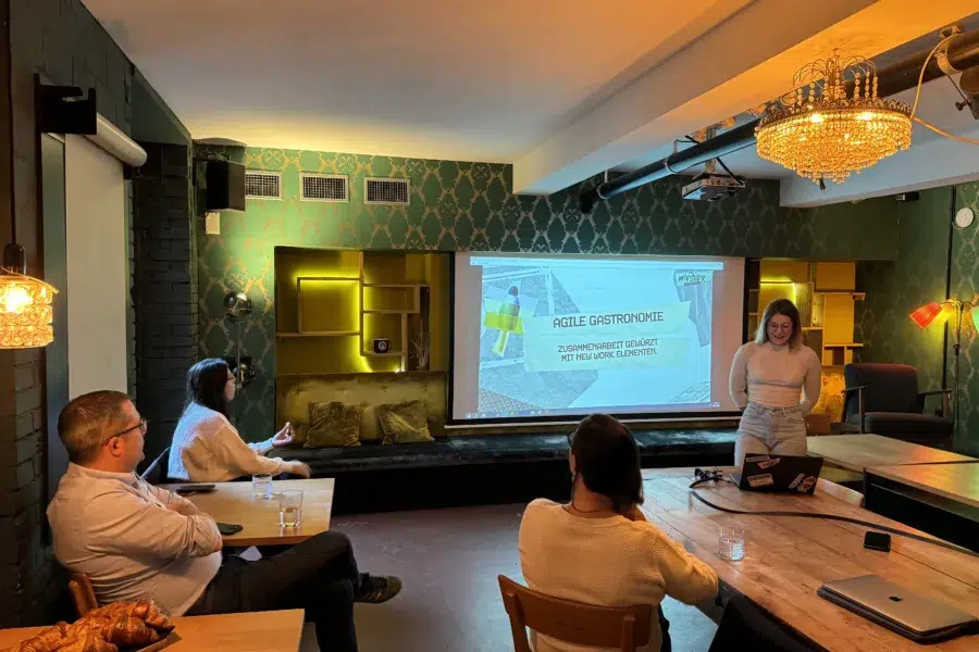Eine Frau präsentiert einer kleinen Gruppe in einem stilvollen Raum eine Diashow mit dem Titel „Agile Gastronomie“ und erkundet dabei die Synergie von Küche und neuen Arbeitsmethoden. Die Teilnehmer sitzen mit Laptops und Gebäck um einen Holztisch inmitten warmer Tapeten, eines Kronleuchters und stimmungsvoller Beleuchtung.