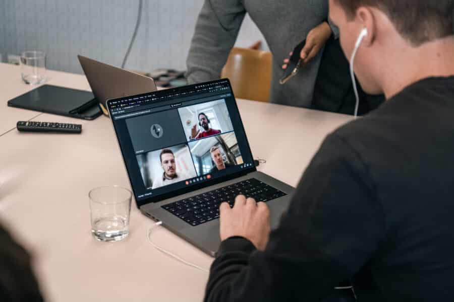 Eine Person mit Kopfhörern nimmt an einer Videokonferenz von Panter News über einen Laptop teil, auf dem Bildschirm sind drei weitere Personen zu sehen. In der Nähe steht jemand mit einem Telefon. Auf dem Tisch stehen ein Laptop und ein Glas Wasser.