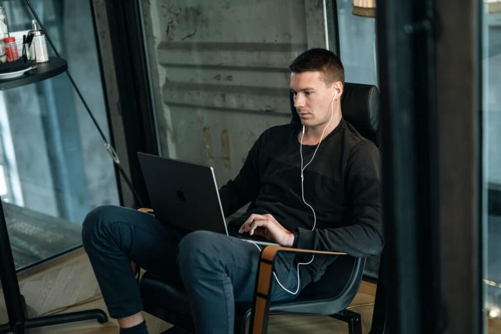 Ein Mann sitzt in einem modernen Stuhl neben einem Fenster und benutzt einen Laptop. Er trägt ein dunkles Sweatshirt, eine dunkle Hose und Kopfhörer und scheint sich auf seine Arbeit zu konzentrieren. In der Nähe ist ein kleiner runder Tisch mit Gewürzen und einer Flasche zu sehen. Die Umgebung wirkt lässig und modern. Ein Mann sitzt in einem modernen Stuhl neben einem Fenster und benutzt einen Laptop. Er trägt ein dunkles Sweatshirt, eine dunkle Hose und Kopfhörer und scheint sich auf seine Arbeit zu konzentrieren. In der Nähe ist ein kleiner runder Tisch mit Gewürzen und einer Flasche zu sehen. Die Umgebung wirkt lässig und modern.