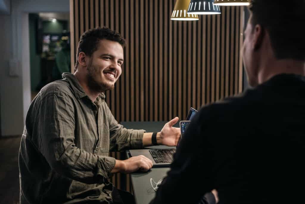 Ein Mann mit kurzen lockigen Haaren und Bart, der ein grünes Hemd trägt, lächelt, während er mit einem Laptop an einem Tisch sitzt. In einer modernen Büroumgebung gestikuliert er mit seiner linken Hand in Richtung einer anderen Person, deren Rücken der Kamera zugewandt ist. Ein Mann mit kurzen lockigen Haaren und Bart, der ein grünes Hemd trägt, lächelt, während er mit einem Laptop an einem Tisch sitzt. In einer modernen Büroumgebung gestikuliert er mit seiner linken Hand in Richtung einer anderen Person, deren Rücken der Kamera zugewandt ist.