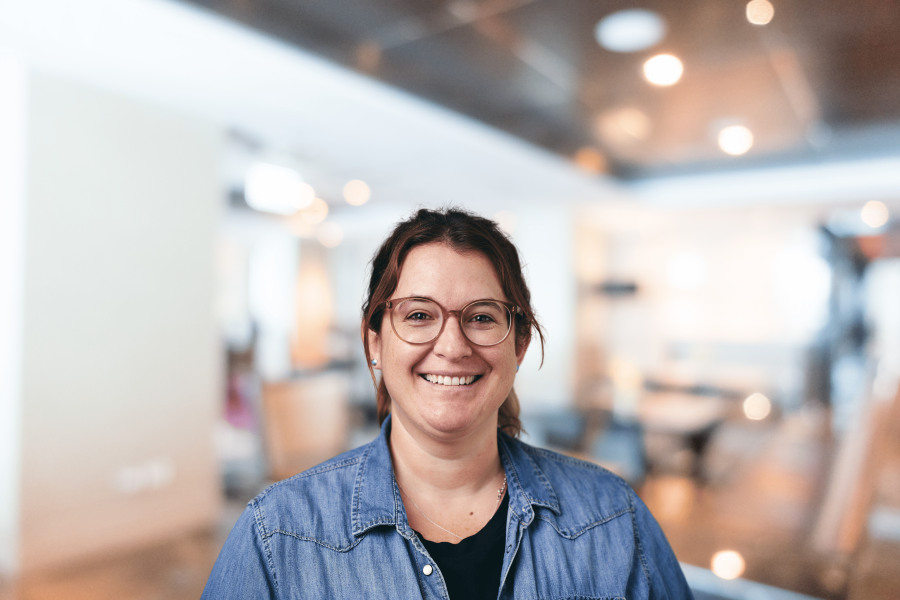 A person with glasses is smiling while standing in a brightly lit indoor setting, embodying the spirit of community building. They wear a denim jacket over a black top, radiating warmth and inclusivity against the softly blurred background.