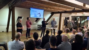 Vier Männer stehen vor einem modernen Loft mit freiliegenden Balken und halten eine Präsentation vor einem sitzenden Publikum. Eine Folie mit dem Titel "Is SaaS Dead?", auf der ein Roboterarm abgebildet ist, wird auf dem Bildschirm dieser AI Events-Veranstaltung angezeigt.