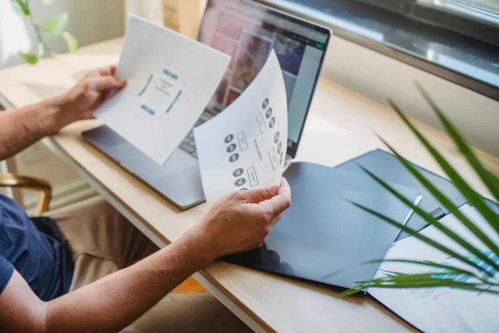 A person reviews printed fintech diagrams in front of a laptop on a wooden desk, with an open black folder nearby. Lush green plant leaves frame the scene, and soft light filters through the window in the background.