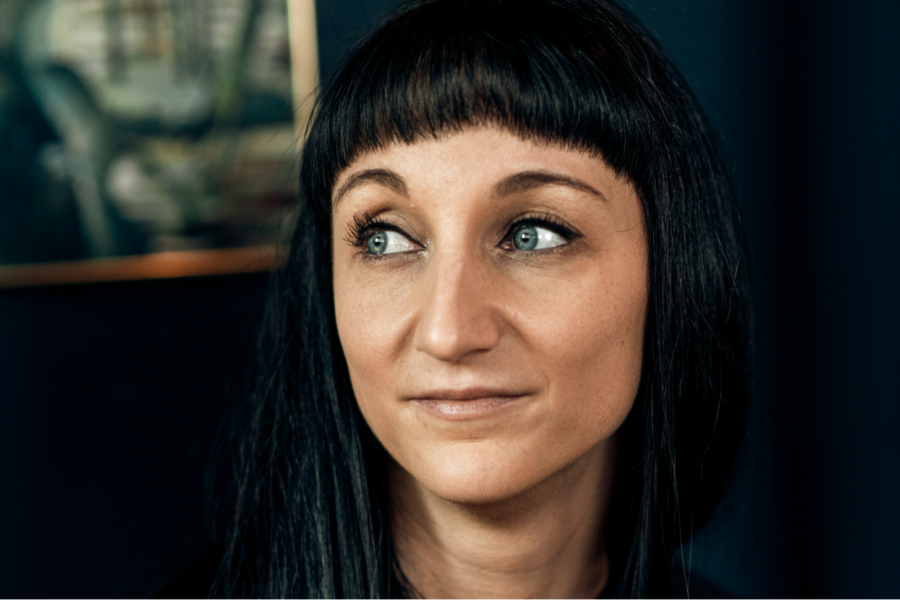 Mit langen schwarzen Haaren und Pony blickt ein UX/UI-Designer nachdenklich zur Seite. Seine blauen Augen und sein subtiles Lächeln deuten auf Kreativität hin, während im dunklen Hintergrund ein undeutliches Kunstwerk auftaucht.