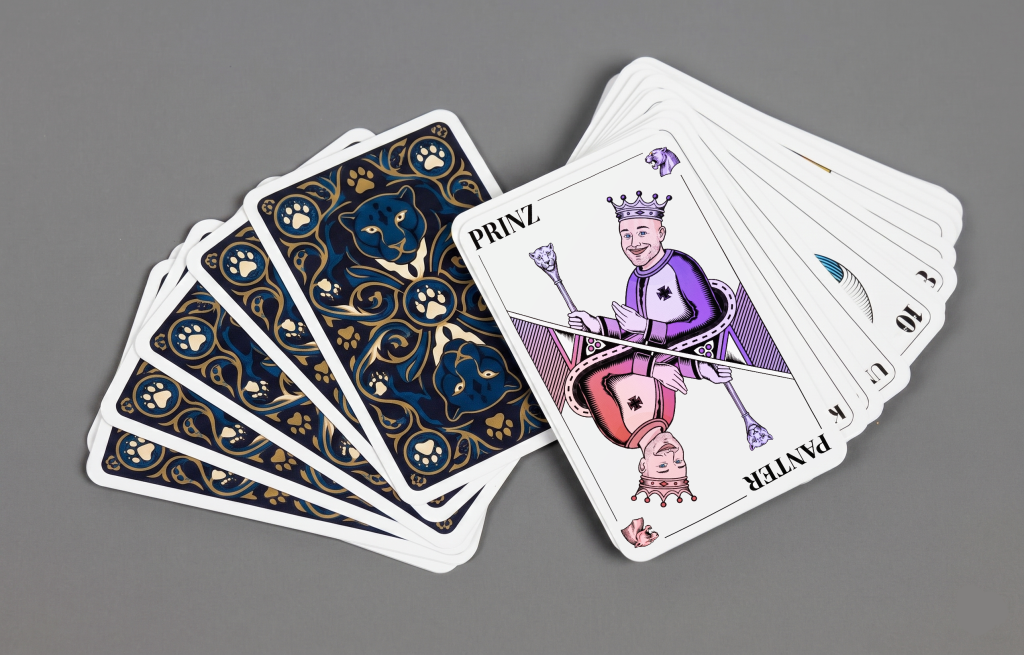A deck of cards fanned out on a grey surface, with blue-and-gold lion motifs on the backs — perfect for AI events. On the front, a king is shown, referred to as “Prinz Panter,” wearing purple and red clothing.