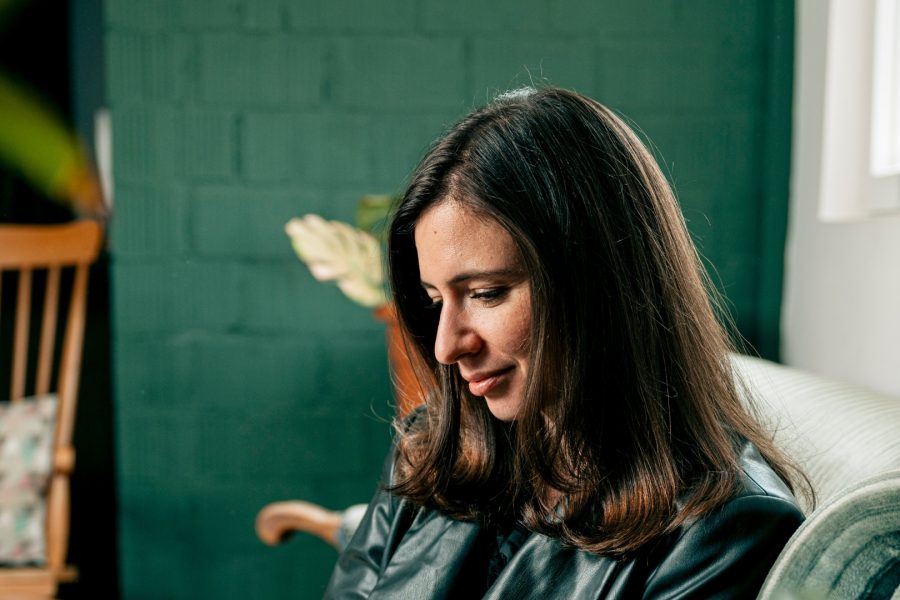 A UX/UI designer with long dark hair and a leather jacket sits on a sofa, looking downward with a gentle smile. In the softly lit room with teal walls, a wooden chair and a potted plant add to the serene atmosphere.