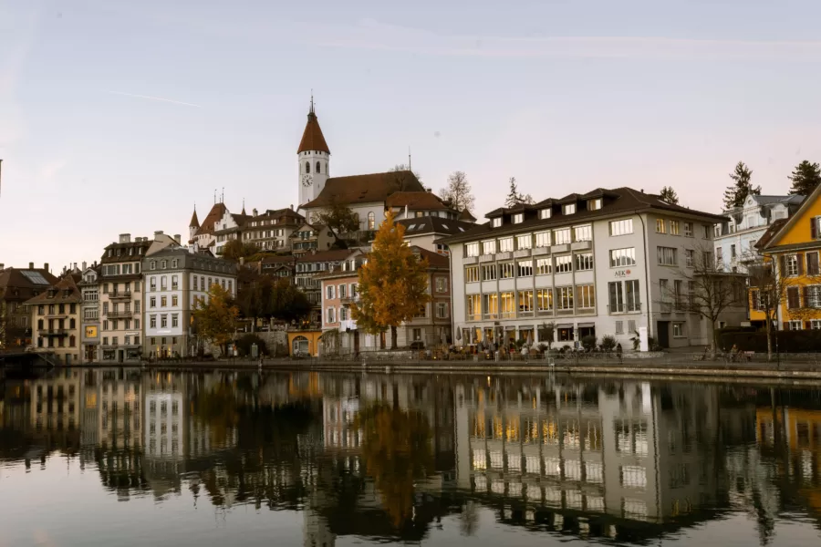 A scenic view of a town with historic buildings by a calm river captures the architecture perfectly, like a well-crafted webseite. A church tower rises prominently in the background, complemented by autumn trees that add vibrant color under the clear sky, hinting at tranquility.