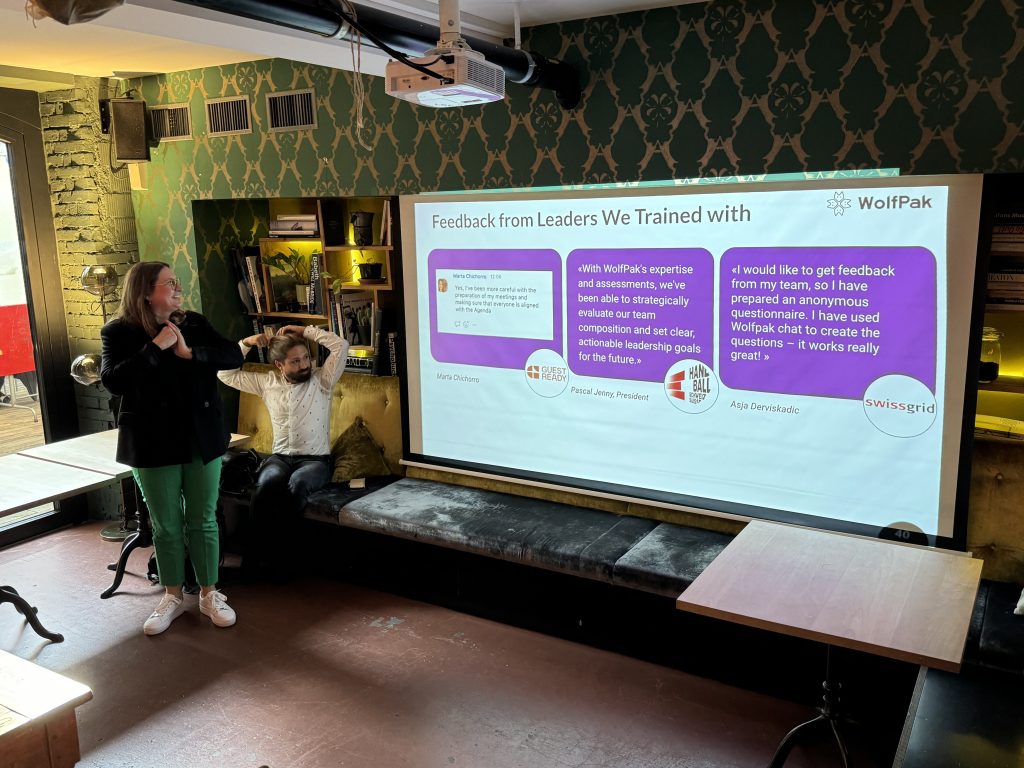 In a dimly lit room, with a modern, casual vibe, a woman confidently presents in front of a screen showcasing WolfPak testimonials during pan.talk. Positive feedback from leaders flashes by as she speaks, while a man sits relaxed on a bench.