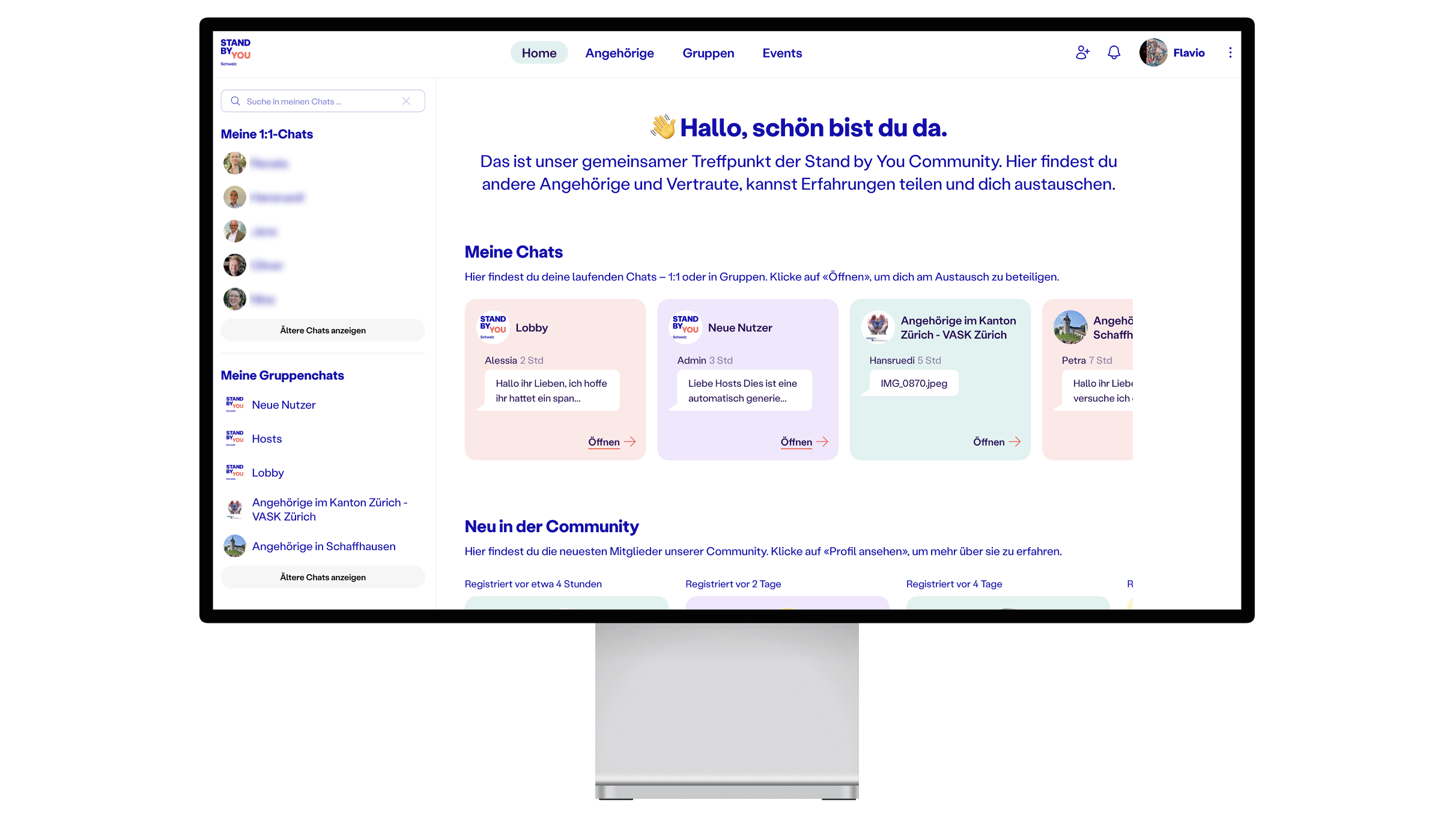 A computer monitor displays a German-language online community and volunteer platform, showing chats, groups, and community updates with a blue and white user interface. A computer monitor displays a German-language online community and volunteer platform, showing chats, groups, and community updates with a blue and white user interface.