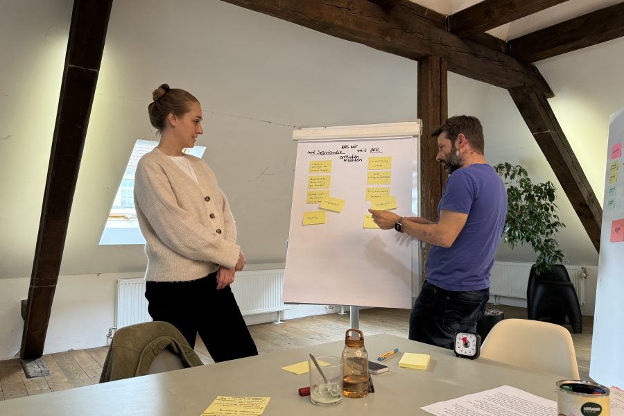 In einer Demonstration von Soziokratie in Aktion unterhalten sich zwei Personen in einem Besprechungsraum über Notizen auf einem Flipchart. Die Frau links trägt einen leichten Pullover und Jeans, während der Mann rechts ein blaues Hemd und Jeans trägt und auf Haftnotizen zeigt, auf denen ihre gemeinsamen Ideen skizziert sind.