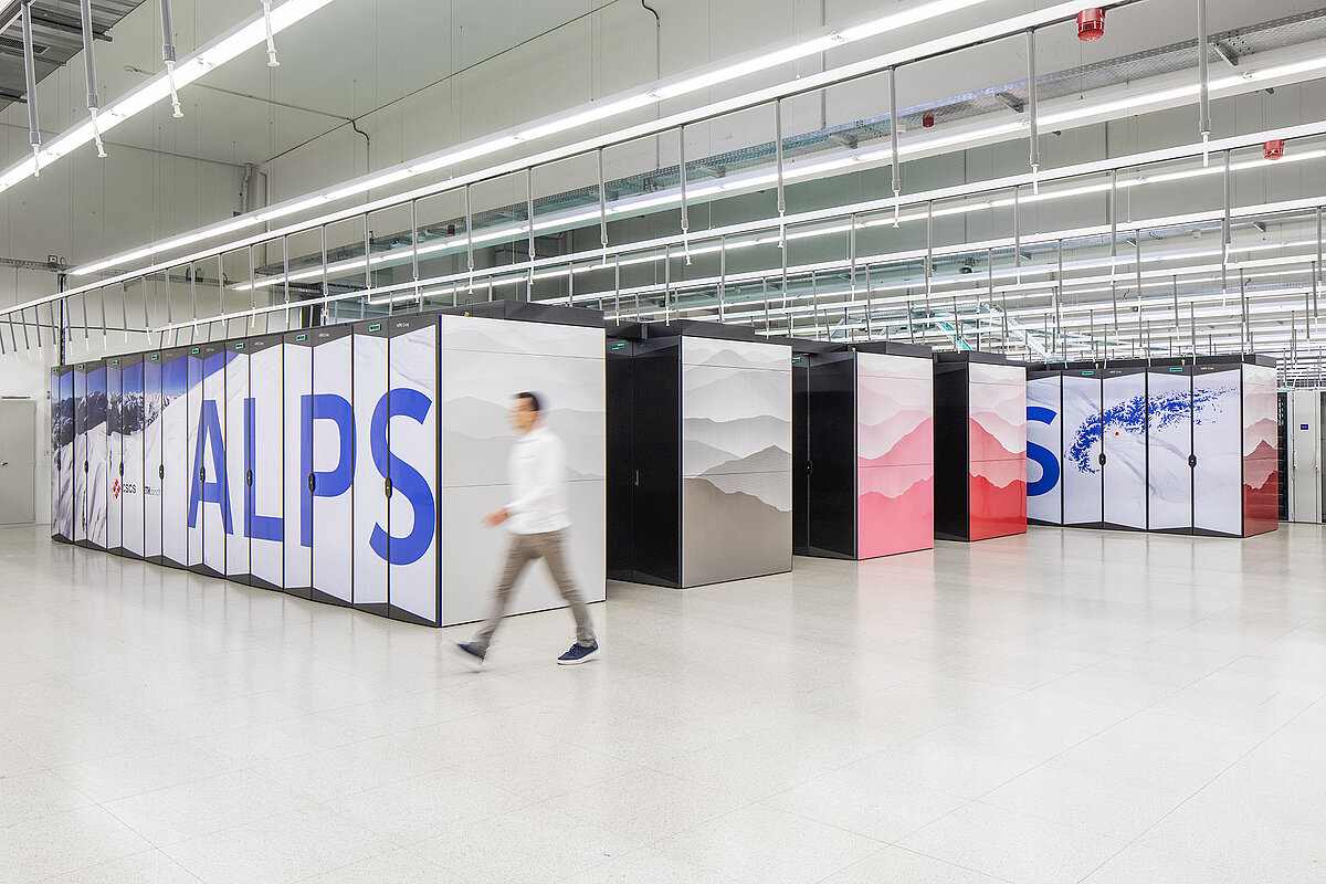 Eine Person geht an großen, modernen Supercomputern in einem hell erleuchteten, großen Raum vorbei. Die Geräte, die zur Schweizer KI-Initiative gehören, sind künstlerisch gestaltet, mit dem Wort ALPS in großen blauen Buchstaben und einem Kunstwerk mit Bergmotiven.