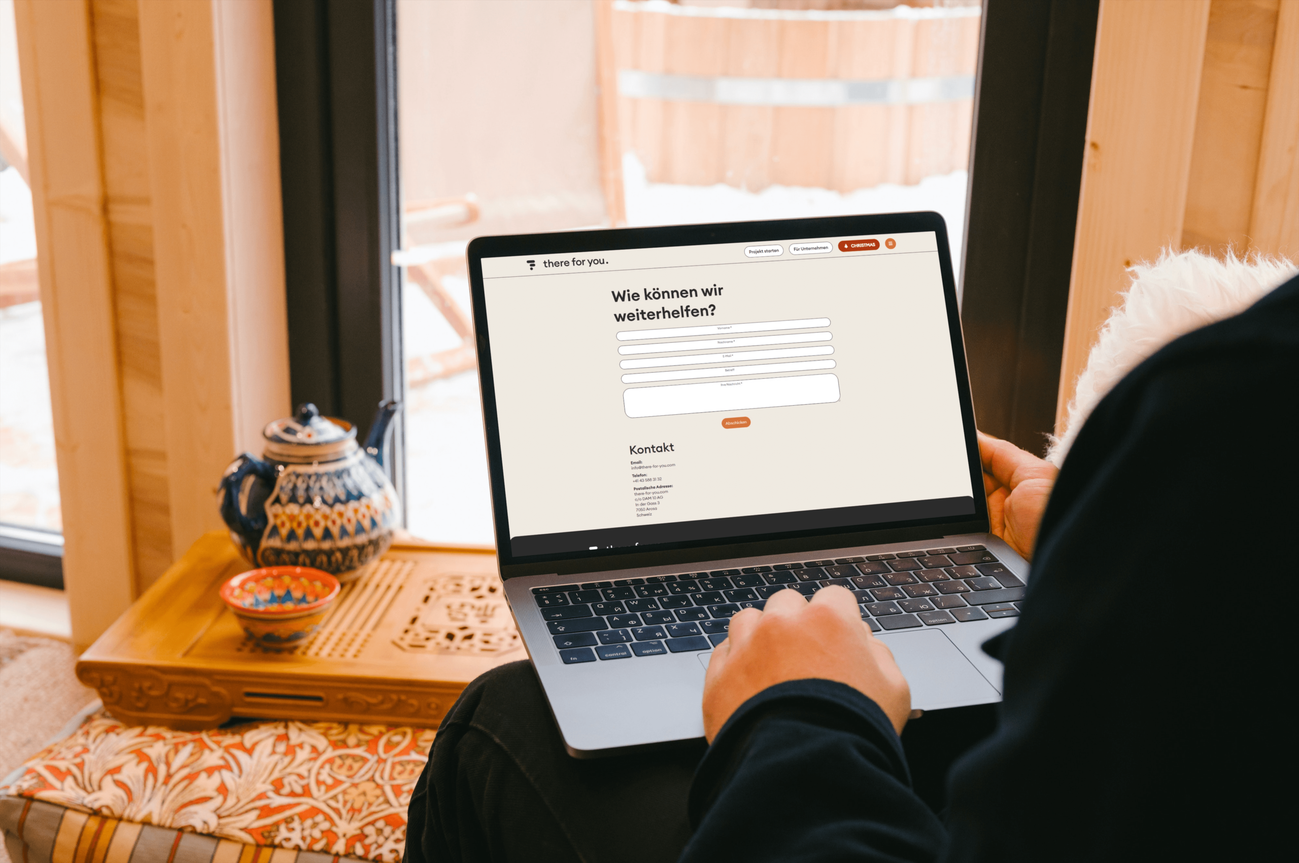 Eine Person sitzt am Fenster und vertieft sich in einen Laptop, auf dem ein deutsches Kontaktformular angezeigt wird – eine Szene, die Startup-Inspiration verkörpert. Daneben stehen eine dekorative Teekanne und eine kleine Schüssel auf einem Holztablett. Licht durchflutet die gemütliche Umgebung und hebt das charmante Holzinterieur hervor.